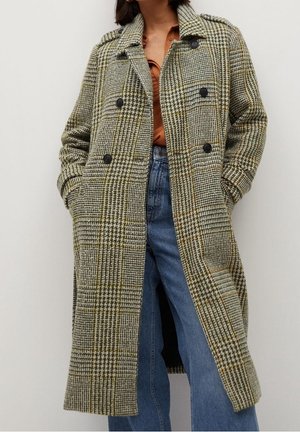 Kvinde iført lang, dobbeltradet jakke med hundetandsmønster over rustfarvet skjorte med knapper og blå jeans, hænderne i kårlommerne.