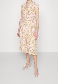 Vestido midi rosa con mangas cortas, que presenta un patrón de cadenas doradas; cinturón en la cintura, falda fluida y combinado con zapatos de tacón cuadrado rosa claro.