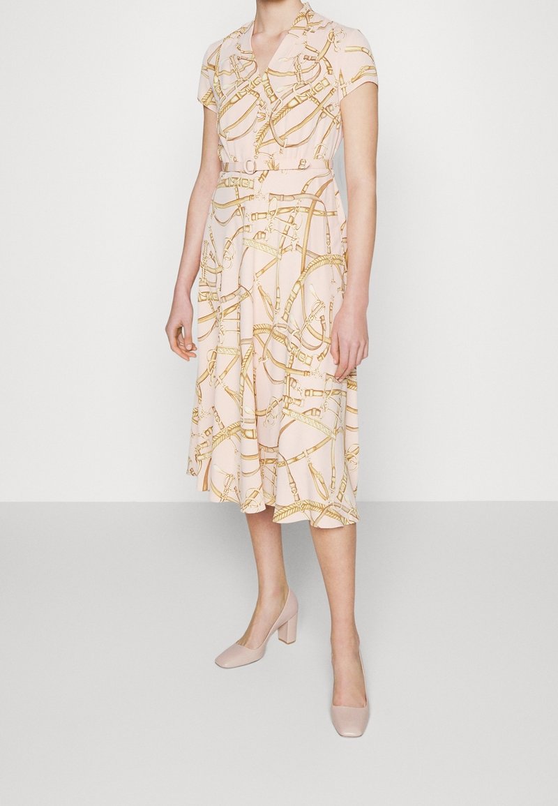 Vestido midi rosa con mangas cortas, que presenta un patrón de cadenas doradas; cinturón en la cintura, falda fluida y combinado con zapatos de tacón cuadrado rosa claro.