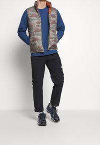 Uomo che indossa un gilet imbottito mimetico, una camicia a maniche lunghe blu, pantaloni cargo neri, calzini grigi e scarpe da trekking nere, in piedi con le mani nelle tasche.