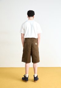 Pantaloni scurți maro tip cargo, cu buzunare mari la spate, purtați cu un tricou alb. Persoana poartă, de asemenea, cizme de tobă bleumarin cu sole ușoare.