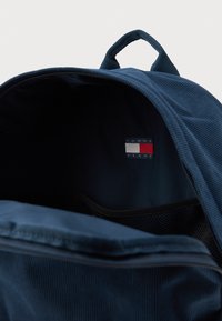 Mørkebl�å rygsæk med et corduroy-yderlag, der har en mesh-lomme og et mærketikette i rød, hvid og blå indeni.