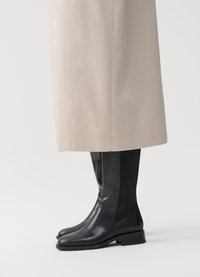 Botas de couro pretas até ao joelho, com um acabamento suave, bico arredondado e painéis laterais elásticos, combinadas com uma peça exterior de cor bege clara.