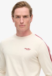 Lange mouwen crèmekleurig T-shirt gemaakt van katoen, met rode strepen op de schouders en een klein geborduurd logo op de borst.