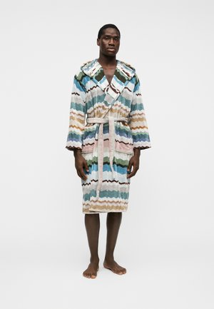 Homme debout, pieds nus, portant un peignoir à capuche avec des rayures en zigzag multicolores et une ceinture nouée, sur un fond blanc uni.
