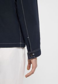 Tamsiai mėlynas švarkas su struktūriniu apykakliu, matomomis baltomis siūlėmis ir sagos detalė ant rankogalių, derinamas su šviesios spalvos kelnėmis.