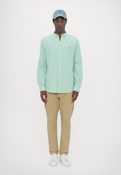 Chemise verte clair à manches longues avec col boutonné, pantalons cargo beiges, casquette bleue et baskets blanches. Design simple sans motifs.