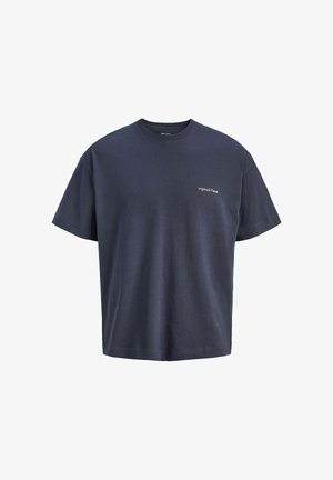 Junge trägt ein dunkelblaues T-Shirt, ausgewaschene Jeans mit weitem Bein und weiße Sneaker, schaut nach rechts vor einfachem Hintergrund.