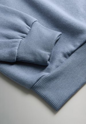 Primer plano de los puños de un sudadera azul claro que muestra la textura de punto acanalado en las mangas y los dobladillos de la cintura sobre una superficie plana.
