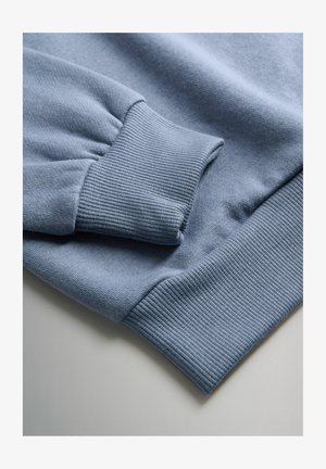 Prim-plan al manșetelor unui hanorac light blue, evidențiind textura tricotată în rind, pe marginile mânecilor și ale taliei, așezat pe o suprafață plană.