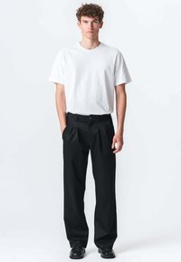 Hvid bomulds T-shirt sat sammen med sorte plisserede bukser. Bukserne har knaplukning og bred ben-design; modellen står lige.