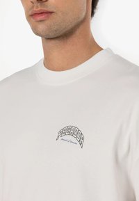 White cotton t-shirt featuring a round neckline and a small embroidered logo that reads "PEGADOR" along with "pursuit of dreams" in blue.