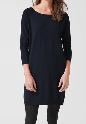 Femme portant une robe noire à manches longues, longueur genou, avec un décolleté en U et des collants noirs, debout devant un fond clair uni.