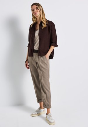 Dark brown button-up shirt, beige trousers with a subtle pattern, and white sneakers. Rolled sleeves and hands in pockets.