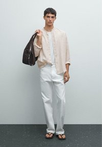 Helles beiges Hemd mit Knopfleiste, weißes T-Shirt, weiße lässige Hose, braune Tasche mit einem Riemen und Sandalen mit schwarzen Riemen. Schlichter Hintergrund.