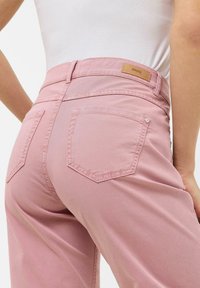 Pantalons ajustés roses avec une texture lisse, comportant deux poches arrière, une taille cousue et une étiquette de marque à l'arrière.