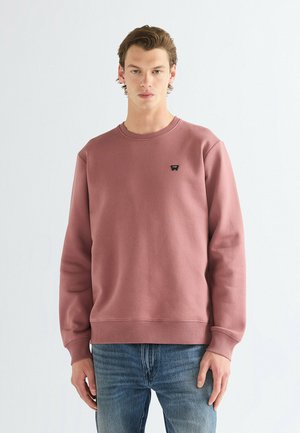 Jeune homme portant un sweat-shirt rose poudré avec un petit logo noir sur la poitrine et un jean bleu, debout devant un fond blanc uni.