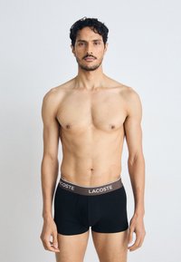 Boxer-briefs noirs avec une ceinture élastique marron arborant le logo "LACOSTE". Tissu lisse, design ajusté et sans motifs ni accents visibles.
