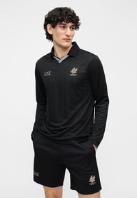 Polo de manga larga negro con tapeta, con un acento azul claro y marca dorada; combinado con pantalones cortos negros, ambos de textura lisa.