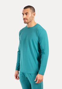 Türkises Langarmshirt aus glattem, leichtem Stoff. Mit einem Rundhalsausschnitt und einer enganliegenden Silhouette, kombiniert mit passenden Hosen.