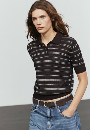 Jong persoon met lang haar, gekleed in een zwart-wit gestreept poloshirt, blauwe spijkerbroek en een bruine riem, lopend met één hand in de pocket.