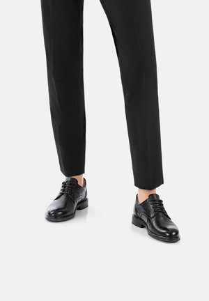 Schwarze Lederschuhe mit runden Zehen, Schnürdesign, strukturiertem Obermaterial und niedrigen Blockabsätzen, getragen mit maßgeschneiderten schwarzen Hosen.