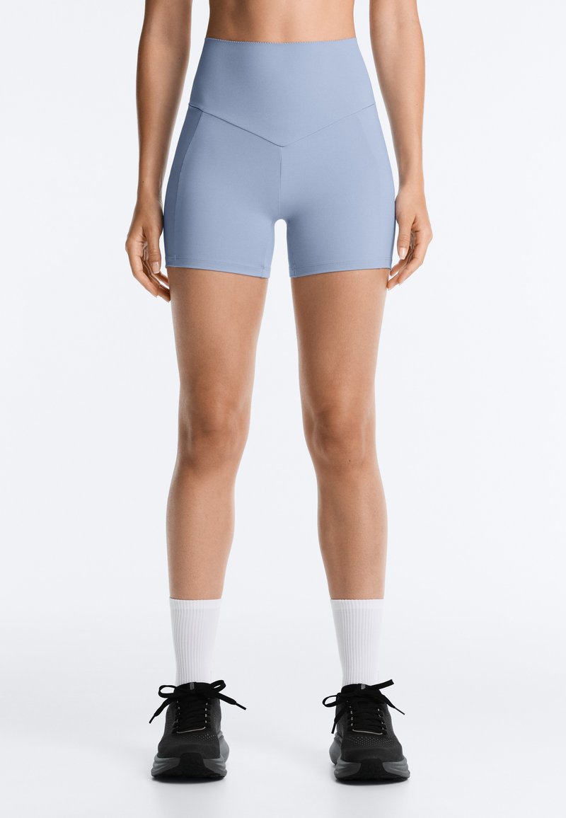 Femme portant un short ajusté taille haute bleu clair, des chaussettes blanches et des chaussures de sport noires, debout sur un fond clair uni.