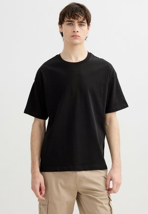 Joven con cabello castaño, vestido con una camiseta negra lisa y pantalones cargo beige, sobre un fondo claro y liso.