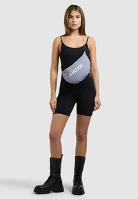 Junge Frau in einem schwarzen ärmellosen Overall und schwarzen Kampfboots, die eine graue Umhängetasche mit der Aufschrift "KHUJO" trägt.