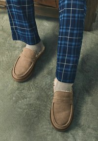 Osoba oblečená v béžových zamšových mokasínach, svetlobéžových ponožkách a modrých kockovaných nohaviciach stojí na sivej koberci pri drevenom nábytku.