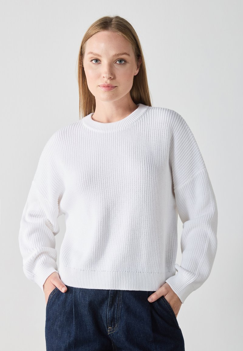 Pull en maille côtelée blanche avec un col rond et une coupe légèrement oversized. Dispose de manches longues et d'un tissu doux et texturé.