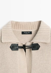 Beige knitted cardigan with a ribbed collar. Features a black toggle closure and textured knit pattern. Label visible at the neck.