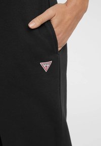 Zwarte joggingsbroek van textuurstof, met een zijzak en een rood-wit driehoekig "GUESS" logo op de linker bovenbeen.