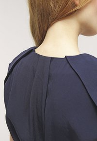 Tmavě modré šaty s záhybovým detailem na zádech, s hladkou texturou a strukturovaným designem. Disponují zaobleným výstřihem.