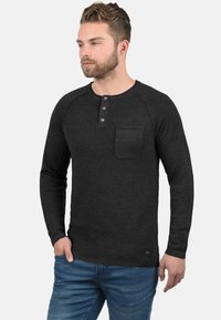 Tamno siva long-sleeve henley majica s teksturiranim materijalom, placketom s tri gumba i džepom na prsima. Uparena s plavim trapericama.