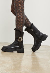 Botas negras hasta el tobillo con un acabado de cuero brillante, que cuentan con un borde de piel sintética, cierre lateral y una suela gruesa y texturizada. Detalle decorativo de hebilla.