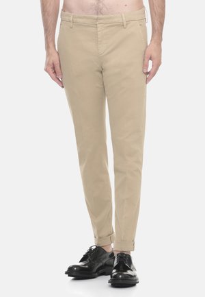 Uomo che indossa chinos slim-fit beige con orli risvoltati e scarpe eleganti in pelle nera, in piedi davanti a uno sfondo bianco semplice.