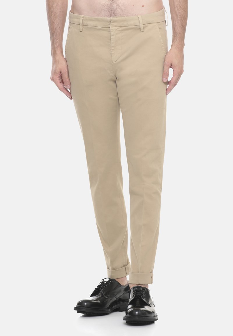 Uomo che indossa chinos slim-fit beige con orli risvoltati e scarpe eleganti in pelle nera, in piedi davanti a uno sfondo bianco semplice.