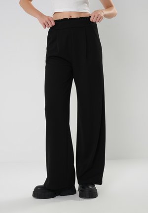 Osoba w czarnych, wysokich, szerokich spodniach i masywnych czarnych butach, regulująca pas spodni na białym, krótkim topie.