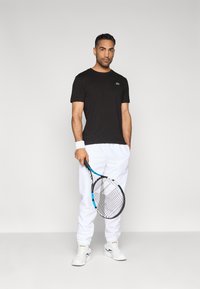 Czarny t-shirt z okrągłym dekoltem z małym logo, zestawiony z białymi spodniami dresowymi i białymi tenisówkami. Trzyma czarno-niebieską rakietę tenisową.