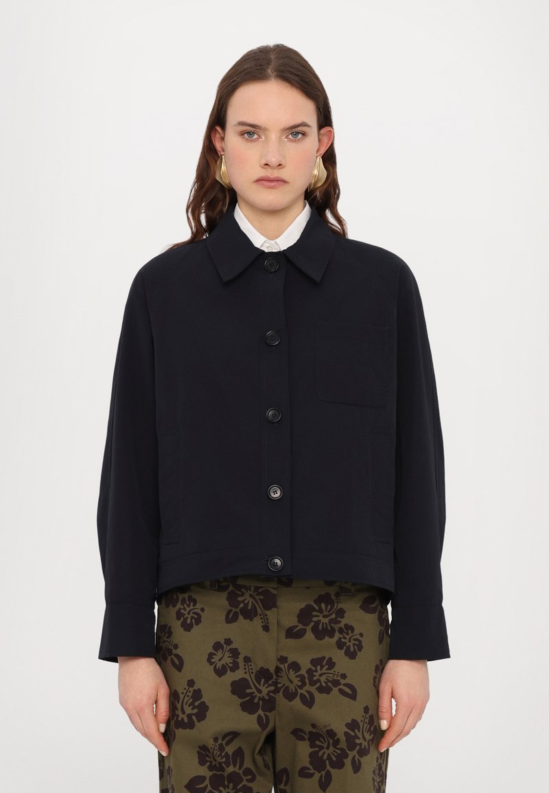 Navyblauwe cropped jas gemaakt van gladde stof, met een kraag, knopen aan de voorkant en een enkele borstzak. Gecombineerd met gestreepte broeken.