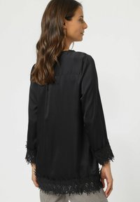 Blusa negra con mangas largas y fluidas, que presenta un borde de encaje en los puños y el dobladillo, con una textura suave y un corte sencillo.