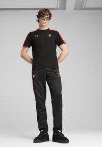 Schwarz kurzärmliges Hemd mit roten Akzenten, Logo auf der Brust; schwarze Trainingshose mit roten Seitenstreifen und Logo; schwarze Wildledersneaker.