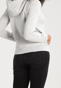 Sudadera gris con capucha, de textura suave y bolsillo tipo canguro, combinada con jeans negros ajustados. El diseño presenta un ajuste casual y relajado.