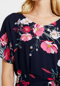 Blumenstickkleid mit navyblauem Hintergrund und großen rosa und weißen Blumen. V-Ausschnitt, kurze Ärmel und eine gebundene Taillenpartie.