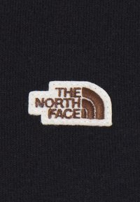 Un tessuto nero presenta una toppa bianca con il logo "THE NORTH FACE" in marrone, con un'icona di montagna curva sul lato sinistro.