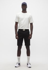 Άντρας που στέκεται φορώντας λευκό πουκάμισο με κοντά μανίκια, μαύρο σορτς με ζώνη, λευκές κάλτσες, λευκά και μαύρα αθλητικά παπούτσια και σκούφο ναυτικού μπλε χρώματος.