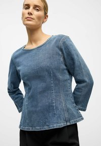 Denimowa bluzka z okrągłym dekoltem, długimi rękawami i delikatnie rozszerzanym dołem. Posiada subtelne detale przeszyć i średni niebieski odcień.
