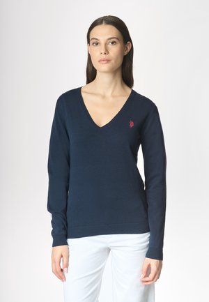 Modella che indossa un maglione blu navy a V con un piccolo logo rosso, abbinato a pantaloni bianchi su uno sfondo chiaro.