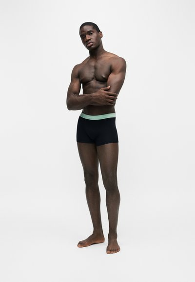 Homme athlétique debout pieds nus portant un boxer noir avec une ceinture vert clair, bras croisés sur la poitrine devant un fond uni.
